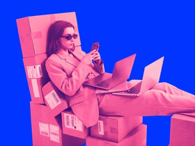 Женщина в костюме сидит на коробках с ноутбуками на коленках, смотрит в телефон.