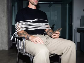 Мужчина, связанный проводом, сидит на стуле и смотрит в телефон