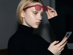 Девушка поднимает розовые очки и смотрит в смартфон