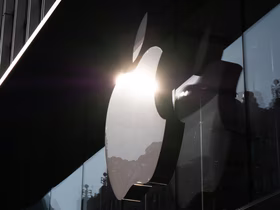 Логотип Apple на стеклянном фасаде фирменного магазина. Сквозь эмблему светит солнце, а в стекле отражаются городские здания и небо