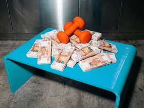 Куча из пачек денег лежит на столе, а гантели прижимают ёё сверху