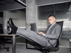 Мужчина в деловом костюме сидит на стуле в офисе, закинув ноги на стол и расположив на коленях ноутбук