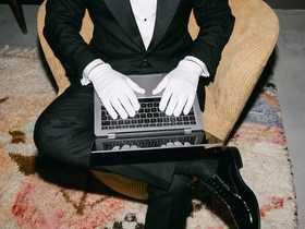 Мужчина в офисном костюме и белых перчатках сидит на пуфе с ноутбуком