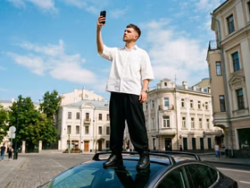 Парень смотрит в телефон, стоя на лобовом стекле машины