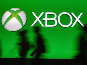 Логотип Xbox — бело-зелёный сферический знак и надпись «XBOX» — на ярко-зелёном фоне; внизу видны размытые силуэты людей