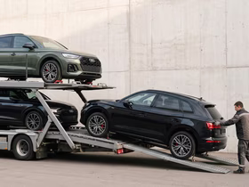 Автовоз с двумя чёрными и зелёными Audi SUV у белой стены, рабочий контролирует процесс