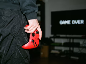 Человек держит в руке красный геймпад Xbox напротив телевизора с надписью Game Over