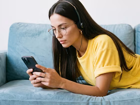 Девушка в очках и наушниках лежит на голубом диване и смотрит в смартфон