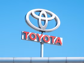 Логотип Toyota на высокой стеле у автосалона на фоне ясного голубого неба. На белой конструкции размещены серебристый фирменный знак и красная надпись Toyota