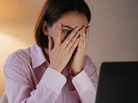 Женщина закрывает лицо руками и смотрит в компьютер
