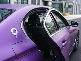 Фиолетовый автомобиль с открытой задней дверью, припаркованный у современного здания. На кузове виден логотип в виде буквы «Q»