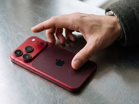 Рука тянется к красному смартфону Apple, лежащему на металлической поверхности; на задней панели видны три камеры.