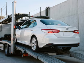 Белый седан Toyota Camry на нижней платформе автовоза; автомобиль частично загружен, рядом видны металлические конструкции транспортировочной платформы и бетонная стена