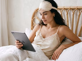 Девушка в бежевой пижаме лежит в постели и смотрит в Ipad