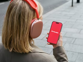 Девушка со светлыми волосами в красных накладных наушниках держит смартфон с красным экраном музыкального приложения; снимок сделан со спины на фоне городской мостовой.