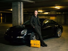 Мужчина опирается спиной на машину, рядом с ним стоит жёлтый портфель