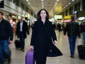 Девушка в чёрном пальто держит в руках фиолетовый чемодан