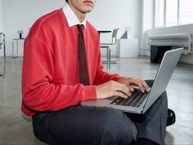 Мужчина в красной деловой рубашке и чёрном галстуке держит на коленях ноутбук