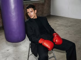 Мужчина в деловом костюме и красных боксёрских перчатках сидит на стуле на фоне фиолетовой боксёрской груши