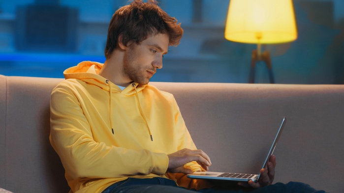 молодой человек, работающий из дома вечером, сидящий на диване с ноутбуком